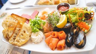 A plate featuring smoked salmon, mussels, fresh vegetables, and lemon, with dipping sauce.