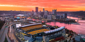 Pittsburgh skyline and Acrisure Stadium glowing at sunset, with the Three Rivers and city landscape creating a stunning aerial view of the Steel City.